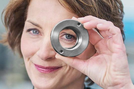 Eine Frau mit kurzen braunen Haaren hält einen metallischen Ring vor ihr rechtes Auge und blickt direkt in die Kamera.